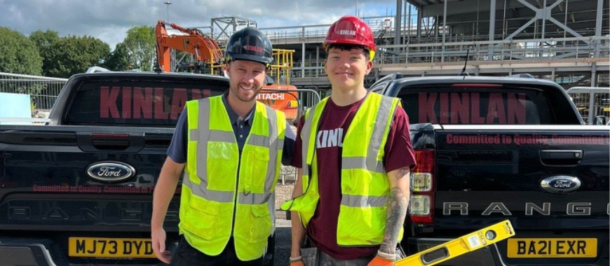 Harry stood with a colleague, wearing a high visibility vest, hard hat and holding a spirit level